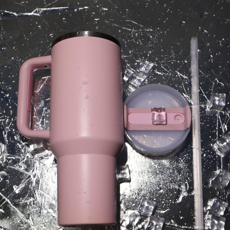 K&ouml;nnte beinhalten: Ein rosa isolierter Becher mit Griff, Deckel und Strohhalm. Der Becher ist zylindrisch mit einem Griff an der Seite. Der Deckel ist hellrosa mit einem transparenten Kunststoffschieber. Ein klarer Strohhalm liegt neben dem Deckel. Eisw&uuml;rfel sind verstreut.