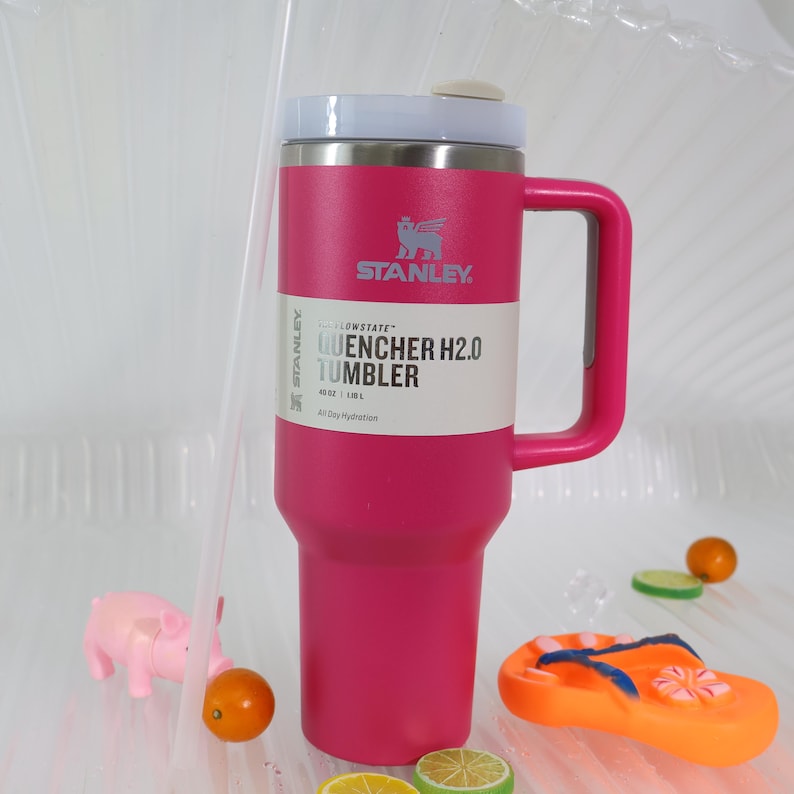 Pu&ograve; includere: Un bicchiere Stanley Quencher H2.0 rosa con coperchio e manico bianchi. Il bicchiere presenta il logo Stanley e il testo "QUENCHER H2.0 TUMBLER" su un'etichetta bianca. Capacit&agrave;: 1,18 litri.