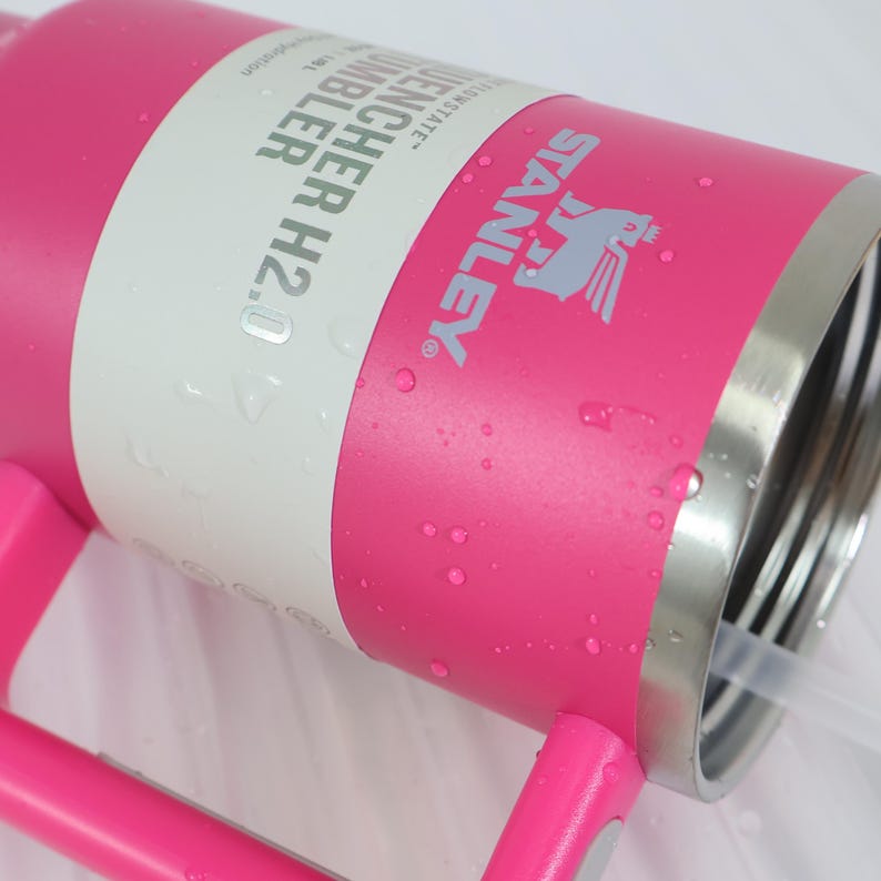 Pu&ograve; includere: Un bicchiere Stanley Quencher H2.0 Flowstate rosa con manico e cannuccia. Il bicchiere presenta il logo Stanley in grigio e le parole "QUENCHER H2.0 TUMBLER" stampate su un'etichetta bianca. Sono visibili goccioline d'acqua.