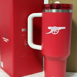 Pu&ograve; includere: Una tazza da viaggio isolante rossa con manico e coperchio bianchi. La tazza presenta il logo dell'Arsenal in bianco e un logo bianco in basso. Una scatola rossa &egrave; visibile sullo sfondo.