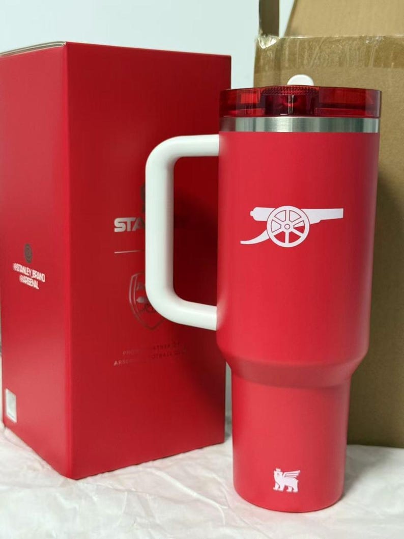 Pu&ograve; includere: Una tazza da viaggio isolante rossa con manico e coperchio bianchi. La tazza presenta il logo dell'Arsenal in bianco e un logo bianco in basso. Una scatola rossa &egrave; visibile sullo sfondo.