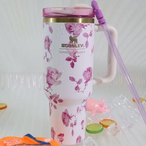 Pode incluir: Caneca isolada branca com um padr&atilde;o floral de rosas cor-de-rosa. A caneca tem uma tampa, al&ccedil;a e canudo cor-de-rosa. O texto "STANLEY" e "LOVESHACKFANCY" est&atilde;o impressos na caneca.