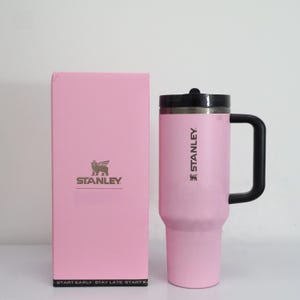 Pu&ograve; includere: Un bicchiere da viaggio Stanley rosa con manico e coperchio neri, accanto a una scatola rosa con il logo Stanley. La tazza ha il marchio Stanley stampato sul lato. La scatola ha il testo "START EARLY. STAY LATE. START B" in basso.