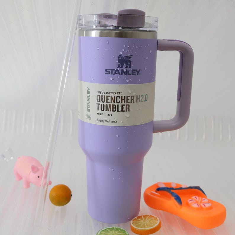 Puede incluir: Un vaso Stanley Quencher H2.0 lavanda con asa, con el logotipo de Stanley y detalles del producto. El vaso es de 1,18 L. La imagen incluye una figura de cerdo rosa, una peque&ntilde;a naranja y una chancla naranja.