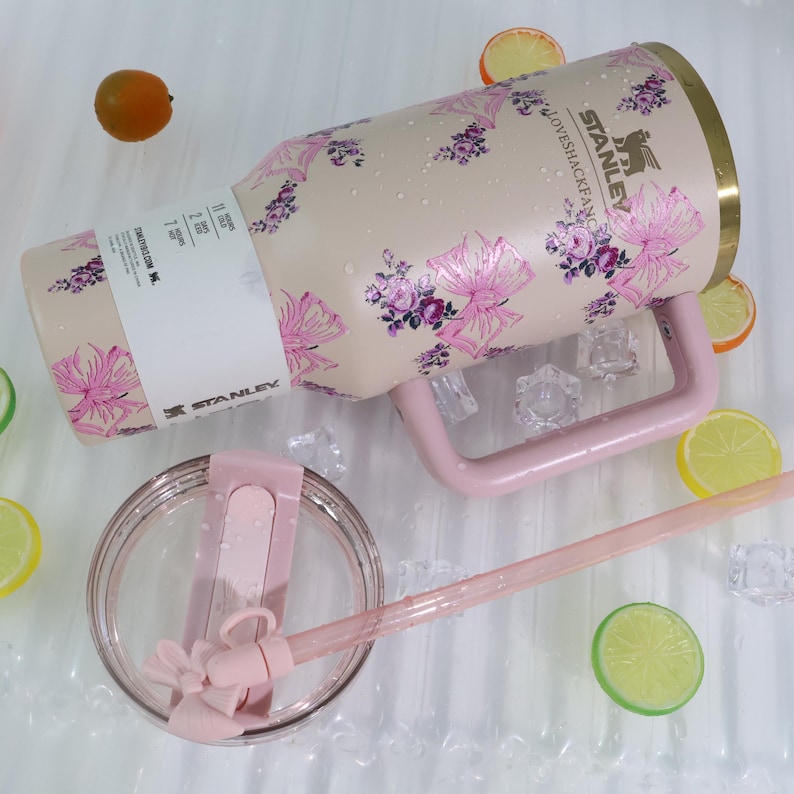 K&ouml;nnte beinhalten: Ein Stanley-Reisebecher mit einem rosa Blumenmuster. Der Becher hat einen rosa Griff und einen transparenten Deckel mit einem rosa Strohhalm. Der Becher ist beige mit rosa und lila Blumen. Auf einer wei&szlig;en Oberfl&auml;che mit Eisw&uuml;rfeln und Zitronenscheiben.