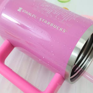 Op de afbeelding: Een roze Stanley + Starbucks-beker met een handvat en een rietje. De beker heeft een zilveren rand en is bedekt met waterdruppels. De tekst "STANLEY + STARBUCKS" staat op de zijkant.