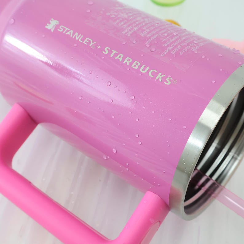 Op de afbeelding: Een roze Stanley + Starbucks-beker met een handvat en een rietje. De beker heeft een zilveren rand en is bedekt met waterdruppels. De tekst "STANLEY + STARBUCKS" staat op de zijkant.