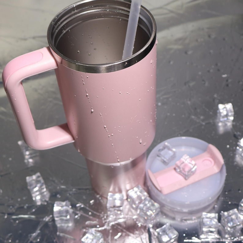 K&ouml;nnte beinhalten: Ein rosa isolierter Becher mit Griff, Edelstahlrand und einem durchsichtigen Strohhalm. Der Deckel ist abgenommen und zeigt Eisw&uuml;rfel. Der Becher steht auf einer reflektierenden Oberfl&auml;che mit verstreuten Eisw&uuml;rfeln.