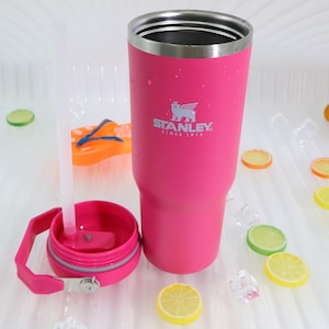 May include: A pink Stanley tumbler with a silver rim and the Stanley logo. The tumbler has a matching pink lid with a handle and a clear straw. The image includes decorative lemon and lime slices, ice cubes, and an orange flip-flop.