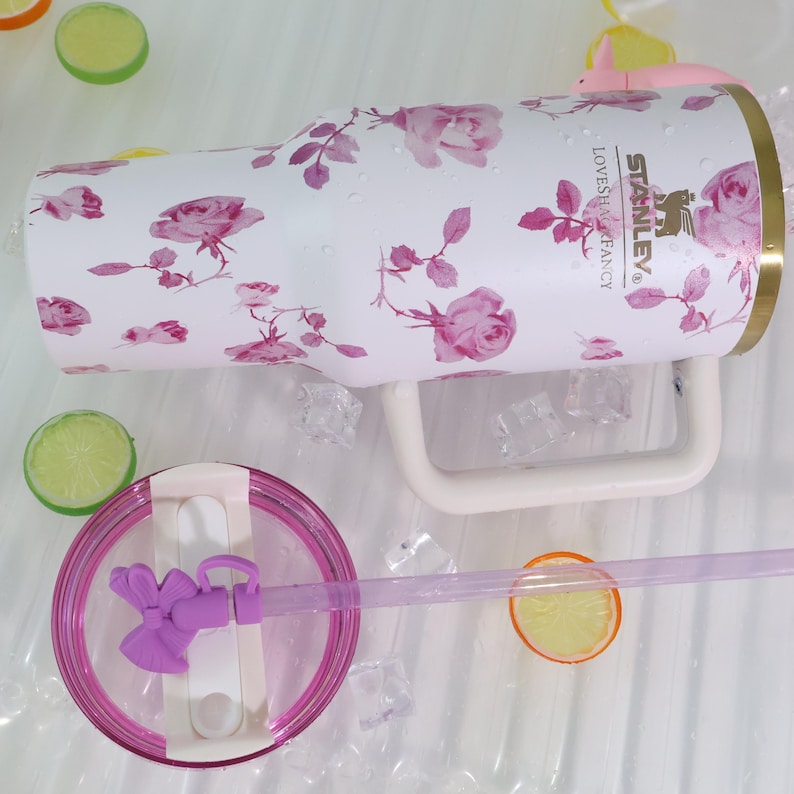 Pode incluir: Caneca isolada branca com um padr&atilde;o floral de rosas em tons de rosa e roxo. A caneca tem uma al&ccedil;a branca e uma tampa transparente com um canudo roxo. O texto "LOVESH&FANCY" e "STANLEY" est&aacute; impresso na lateral.