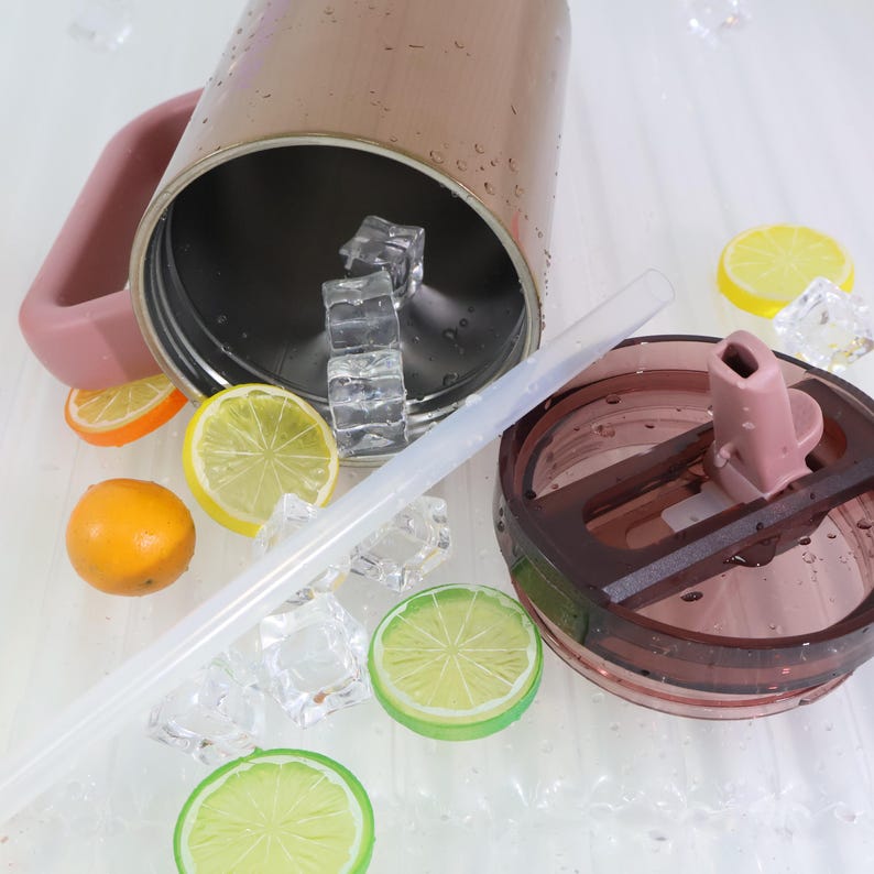 Puede incluir: Un vaso aislado de color oro rosa con un asa y tapa a juego, junto con una pajita transparente. El vaso est&aacute; abierto, revelando cubitos de hielo en el interior. Rodajas decorativas de c&iacute;tricos y m&aacute;s cubitos de hielo est&aacute;n esparcidos alrededor del vaso.