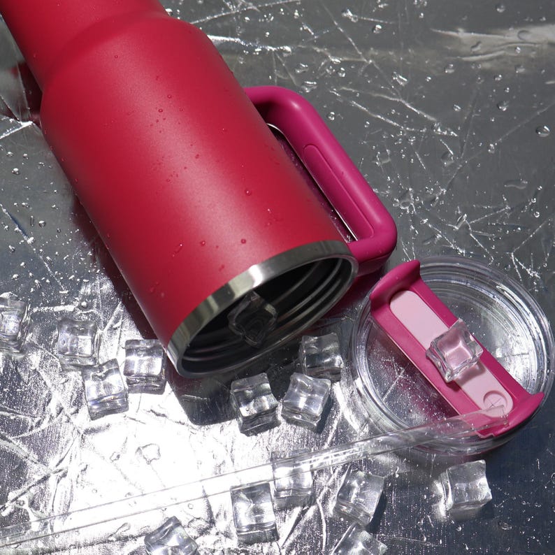 Peut inclure: Un gobelet isotherme rouge avec une poign&eacute;e et un couvercle roses, pos&eacute; sur une surface r&eacute;fl&eacute;chissante avec des gla&ccedil;ons. Le gobelet a un bord argent&eacute; et un couvercle transparent avec un curseur rose. Boissons et hydratation.