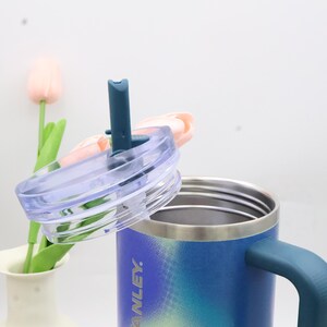 Puede incluir: Una taza de acero inoxidable con degradado azul y verde, tapa transparente y asa turquesa. La tapa tiene una pajita turquesa. La taza tiene la palabra "ANLEY" impresa en el lateral.