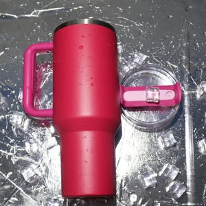 Peut inclure: Un gobelet isotherme rouge vif avec une poign&eacute;e et un couvercle assortis. Le gobelet cylindrique a un bord en acier inoxydable et un couvercle transparent avec un curseur rose. La poign&eacute;e est de forme rectangulaire robuste. Des gla&ccedil;ons entourent le gobelet.