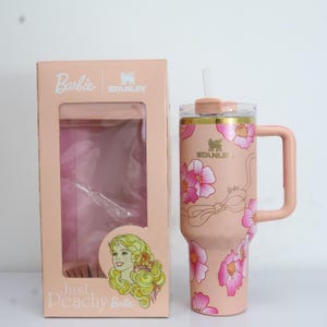 K&ouml;nnte beinhalten: Ein rosa Stanley-Becher mit floralem Design und passendem Griff, neben einer rosa Schachtel mit dem Barbie-Logo. Der Becher hat einen durchsichtigen Strohhalm und einen goldenen Rand. Die Schachtel zeigt eine Barbie-Illustration und den Text "Just Peachy Barbie."