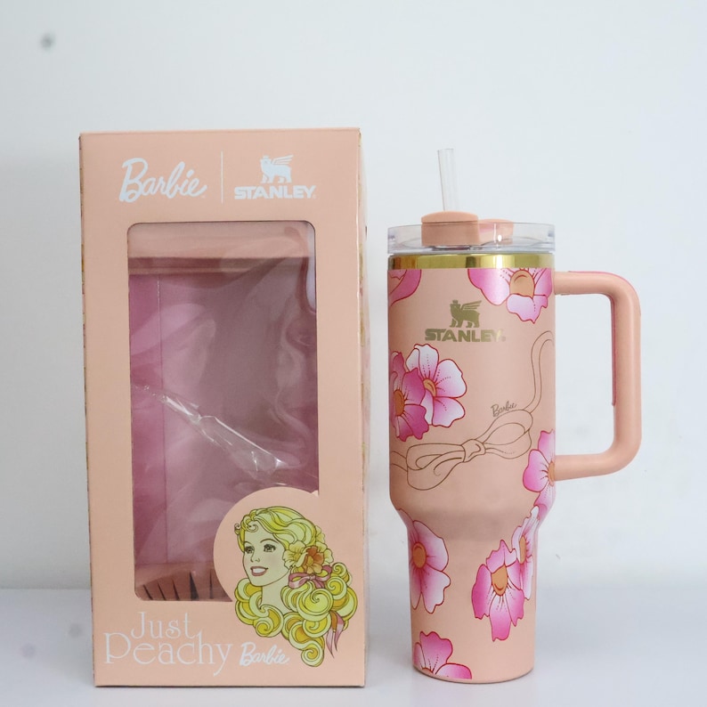 K&ouml;nnte beinhalten: Ein rosa Stanley-Becher mit floralem Design und passendem Griff, neben einer rosa Schachtel mit dem Barbie-Logo. Der Becher hat einen durchsichtigen Strohhalm und einen goldenen Rand. Die Schachtel zeigt eine Barbie-Illustration und den Text "Just Peachy Barbie."