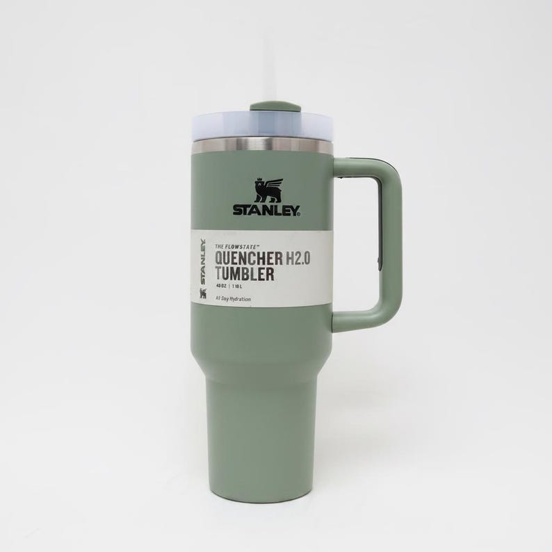 May include: A green Stanley Quencher H2.0 tumbler with a handle and a straw. The tumbler has a white label with black text that reads "QUENCHER H2.0 TUMBLER". The Stanley logo is visible on the tumbler.