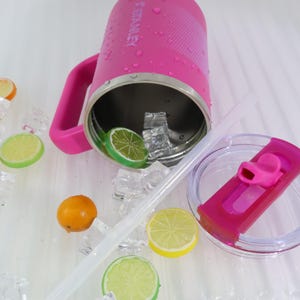 Puede incluir: Un vaso Stanley rosa con asa, tapa y pajita. El vaso est&aacute; parcialmente lleno de hielo y rodajas de lima. La tapa est&aacute; abierta y la pajita est&aacute; sobre la superficie. El vaso tiene la palabra "STANLEY" impresa.