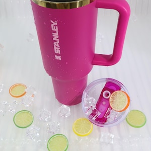 May include: A pink Stanley tumbler with a handle and gold rim, with the Stanley logo. The lid is clear with a pink straw and ice. Decorative lemon and orange slices and ice cubes are scattered around the tumbler.