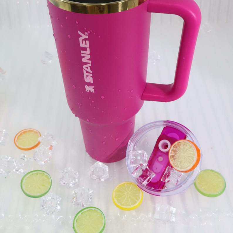 May include: A pink Stanley tumbler with a handle and gold rim, with the Stanley logo. The lid is clear with a pink straw and ice. Decorative lemon and orange slices and ice cubes are scattered around the tumbler.