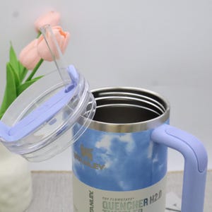May include: A stainless steel tumbler with a light blue and white cloud pattern. It features a light purple handle and lid, and a clear straw. The lid is ajar, showing the inside of the tumbler. The text "STANLEY" is visible on the side.