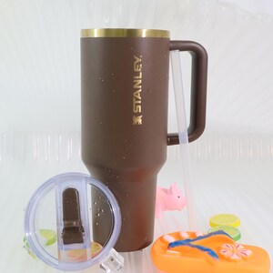 May include: A brown Stanley tumbler with a gold rim and handle, featuring the brand name in gold lettering. A clear lid and straw are included. The image also shows a pink pig, a pair of orange flip-flops, and lemon slices.