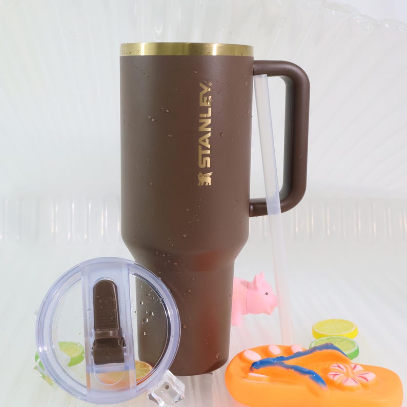 May include: A brown Stanley tumbler with a gold rim and handle, featuring the brand name in gold lettering. A clear lid and straw are included. The image also shows a pink pig, a pair of orange flip-flops, and lemon slices.