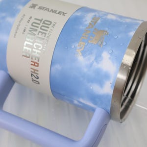 K&ouml;nnte beinhalten: Ein Stanley Quencher H2.0 Becher mit einem hellblauen und wei&szlig;en Wolkenmuster. Der Edelstahlbecher hat einen Griff und einen silbernen Rand. Das Etikett lautet "40 OZ | 1,18 L" und "All Day Hydration".