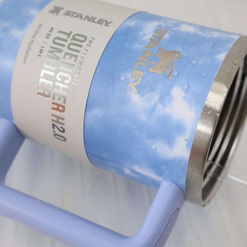 K&ouml;nnte beinhalten: Ein Stanley Quencher H2.0 Becher mit einem hellblauen und wei&szlig;en Wolkenmuster. Der Edelstahlbecher hat einen Griff und einen silbernen Rand. Das Etikett lautet "40 OZ | 1,18 L" und "All Day Hydration".