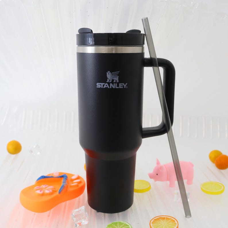 May include: A black Stanley travel mug with a handle and a silver rim. The mug has the Stanley logo and a black lid. A clear straw is next to the mug. The background includes decorative fruit and a pink pig figurine.