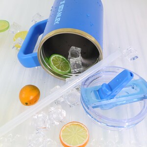 May include: A blue Stanley tumbler with a handle, a clear straw, and a clear lid. The tumbler contains ice cubes and a lime slice. An orange and a lemon slice are also visible.