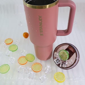 May include: A pink insulated tumbler with a handle and a gold rim, with the brand name "STANLEY" printed on the side. The lid is off, revealing ice cubes and a lime slice. Decorative fruit slices and ice cubes are scattered around the tumbler.