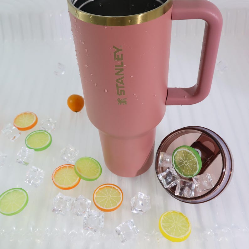 May include: A pink insulated tumbler with a handle and a gold rim, with the brand name "STANLEY" printed on the side. The lid is off, revealing ice cubes and a lime slice. Decorative fruit slices and ice cubes are scattered around the tumbler.
