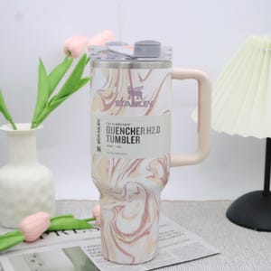 May include: A Stanley Quencher H2.0 Tumbler with a marble design in beige and pink. The tumbler features a handle, a clear lid, and the text "QUENCHER H2.0 TUMBLER". It holds 1.1 L.