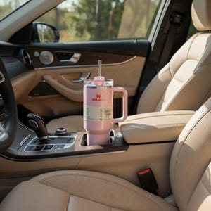 May include: A pink Stanley Quencher tumbler with a handle and straw is in a car cup holder. The tumbler is light pink, with a clear lid and a white label with black text. The car interior has tan leather seats and wood trim.
