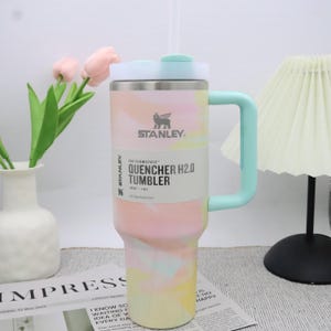 May include: A Stanley Quencher H2.0 tumbler with a pastel tie-dye pattern. The insulated stainless steel tumbler features a mint green handle and lid, and a white straw. The tumbler has the Stanley logo and product information printed on the side.