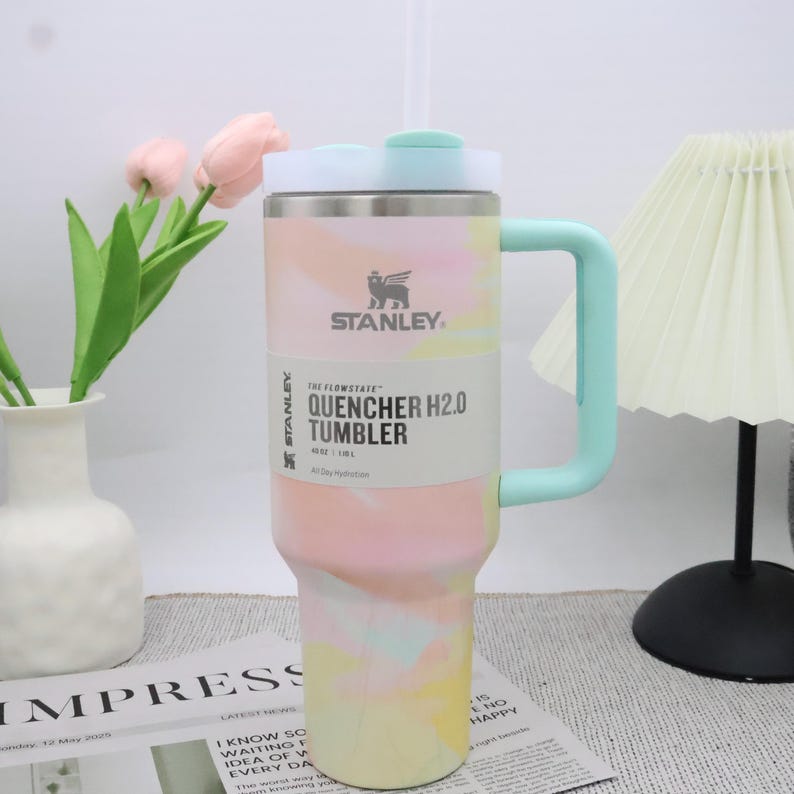 May include: A Stanley Quencher H2.0 tumbler with a pastel tie-dye pattern. The insulated stainless steel tumbler features a mint green handle and lid, and a white straw. The tumbler has the Stanley logo and product information printed on the side.