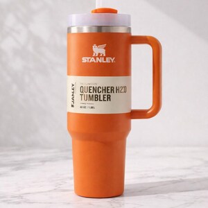 May include: Orange Stanley Quencher H2.0 Tumbler with a white lid, handle, and straw. The tumbler has the Stanley logo and the text "QUENCHER H2.0 TUMBLER" with a capacity of 40 oz / 1.18L. The tumbler is designed for cold beverages.