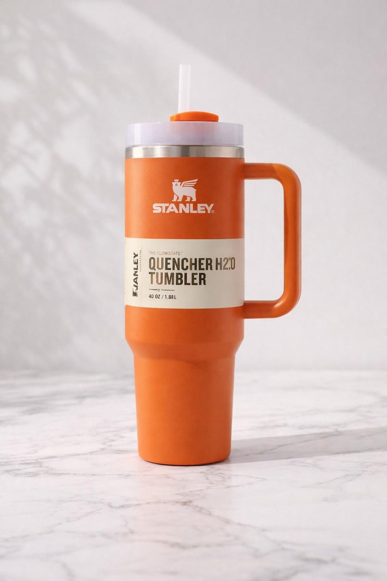 May include: Orange Stanley Quencher H2.0 Tumbler with a white lid, handle, and straw. The tumbler has the Stanley logo and the text "QUENCHER H2.0 TUMBLER" with a capacity of 40 oz / 1.18L. The tumbler is designed for cold beverages.