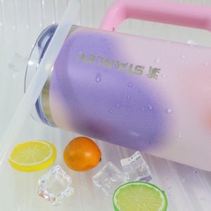 Puede incluir: Una jarra de agua Stanley con un asa rosa y un dise&ntilde;o degradado de morado, rosa y blanco. La jarra tiene una pajita transparente y est&aacute; junto a una rodaja de lim&oacute;n, una naranja, cubitos de hielo y una rodaja de lima.