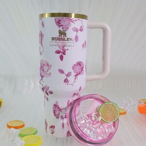 Pode incluir: Uma caneca de viagem Stanley branca com um padr&atilde;o floral de rosas rosa, uma borda dourada e uma al&ccedil;a rosa. A caneca tem o logotipo da Stanley e o texto "LOVESHAL FANCY". A tampa rosa e transparente cont&eacute;m gelo e uma fatia de lim&atilde;o.