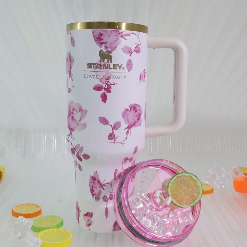 Pode incluir: Uma caneca de viagem Stanley branca com um padr&atilde;o floral de rosas rosa, uma borda dourada e uma al&ccedil;a rosa. A caneca tem o logotipo da Stanley e o texto "LOVESHAL FANCY". A tampa rosa e transparente cont&eacute;m gelo e uma fatia de lim&atilde;o.