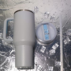 May include: A light gray insulated tumbler with a handle and a lid, next to a clear straw. The tumbler has a silver rim and a light blue accent on the handle. The lid is light gray with a light blue slider. Ice cubes are scattered around the tumbler.