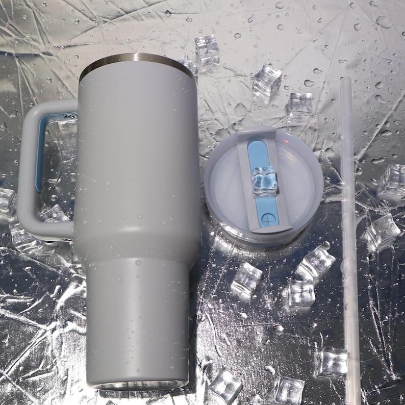May include: A light gray insulated tumbler with a handle and a lid, next to a clear straw. The tumbler has a silver rim and a light blue accent on the handle. The lid is light gray with a light blue slider. Ice cubes are scattered around the tumbler.