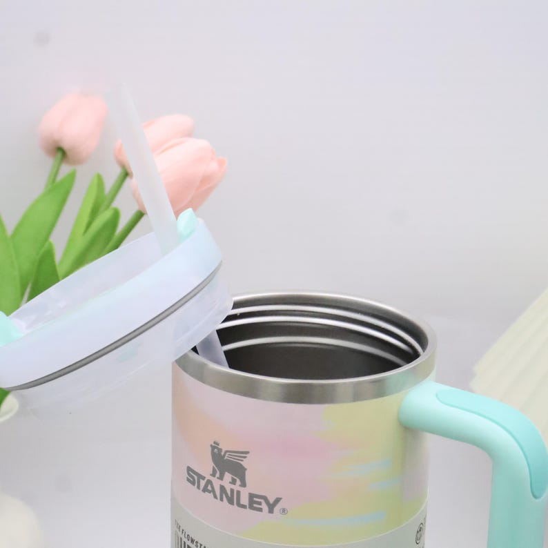 May include: A Stanley insulated mug with a pastel watercolour design in pink, yellow, and blue. The mug has a light blue handle and a matching lid with a straw. The Stanley logo is visible on the front.