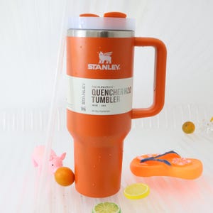 Puede incluir: Un vaso Stanley Quencher H2.0 naranja con tapa y asa blancos y plateados. El vaso tiene el logotipo de Stanley y la informaci&oacute;n del producto impresa. La imagen tambi&eacute;n incluye un cerdito de juguete, una chancla de juguete y fruta.