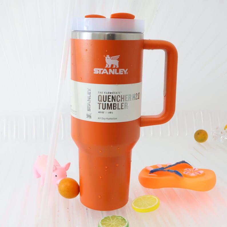 Puede incluir: Un vaso Stanley Quencher H2.0 naranja con tapa y asa blancos y plateados. El vaso tiene el logotipo de Stanley y la informaci&oacute;n del producto impresa. La imagen tambi&eacute;n incluye un cerdito de juguete, una chancla de juguete y fruta.