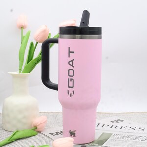 Pu&ograve; includere: Un bicchiere termico rosa con manico e coperchio neri. La parola "GOAT" &egrave; stampata verticalmente in grigio. Un piccolo logo &egrave; visibile in basso. Il bicchiere &egrave; progettato per mantenere le bevande fredde o calde.
