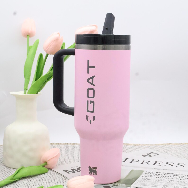 Pu&ograve; includere: Un bicchiere termico rosa con manico e coperchio neri. La parola "GOAT" &egrave; stampata verticalmente in grigio. Un piccolo logo &egrave; visibile in basso. Il bicchiere &egrave; progettato per mantenere le bevande fredde o calde.