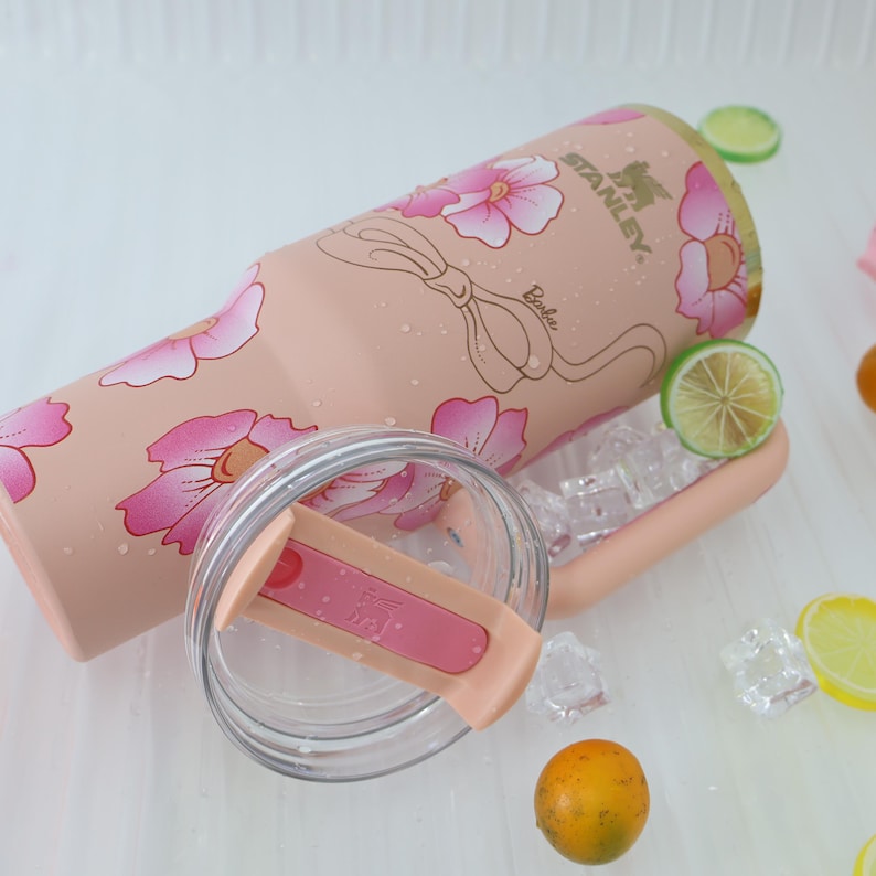K&ouml;nnte beinhalten: Ein rosa Stanley-Becher mit floralem Design und dem Aufdruck "Barbie". Der Becher hat einen transparenten Deckel und ist von Eisw&uuml;rfeln und Zitrusfruchtscheiben umgeben. Der Becher ist f&uuml;r kalte Getr&auml;nke konzipiert.